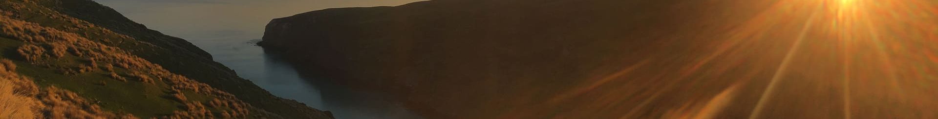 The image depicts a serene sunset over a rugged coastline, with the sun casting vibrant rays and warm light over the landscape. The terrain slopes gently towards a calm sea, and the sky and water are bathed in golden hues, emphasizing the peaceful end to a day in a natural, scenic environment.