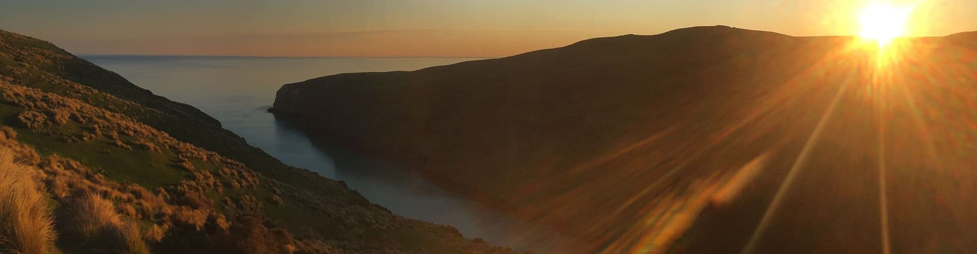 The image depicts a serene sunset over a rugged coastline, with the sun casting vibrant rays and warm light over the landscape. The terrain slopes gently towards a calm sea, and the sky and water are bathed in golden hues, emphasizing the peaceful end to a day in a natural, scenic environment.