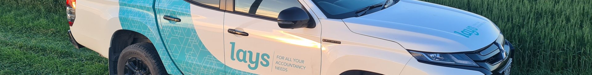 The image features a white pickup truck with blue and turquoise branding for Lay's on its side, parked in a green wheat field during what appears to be early evening. The truck, likely used for promotional or delivery purposes, is shown in a natural, rural setting, emphasizing a connection to farming or agricultural activities.