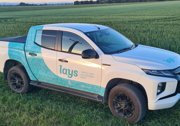 The image features a white pickup truck with blue and turquoise branding for Lay's on its side, parked in a green wheat field during what appears to be early evening. The truck, likely used for promotional or delivery purposes, is shown in a natural, rural setting, emphasizing a connection to farming or agricultural activities.