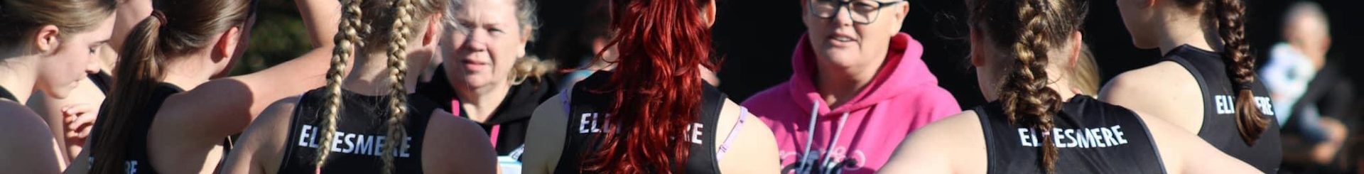 The image shows a group of female netball players in a team huddle, engaging in a discussion with their coach. The coach, wearing a bright pink hoodie, is giving instructions or feedback to the attentive players, who are dressed in matching black and pink uniforms labeled with positions like 'GS' (Goal Shooter) and 'WA' (Wing Attack). This scene likely captures a pre-game or training session moment, emphasizing teamwork and coaching in sports.