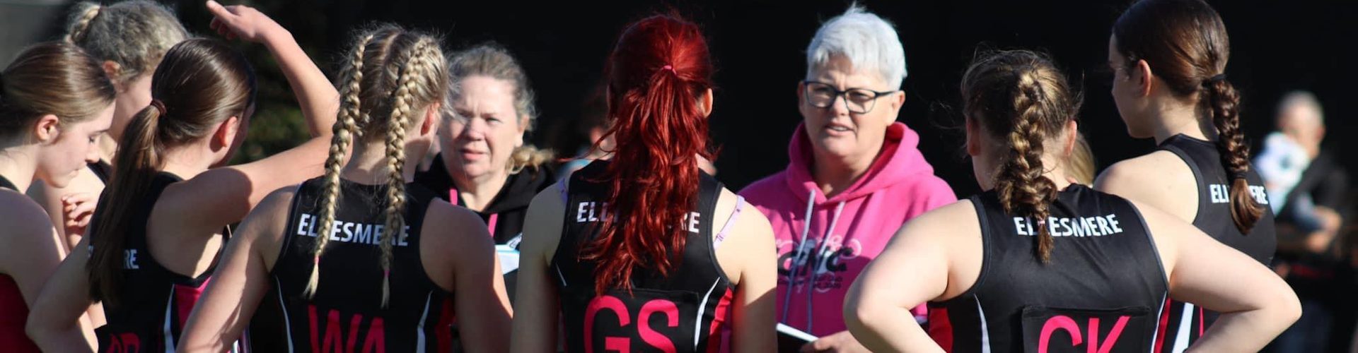 The image shows a group of female netball players in a team huddle, engaging in a discussion with their coach. The coach, wearing a bright pink hoodie, is giving instructions or feedback to the attentive players, who are dressed in matching black and pink uniforms labeled with positions like 'GS' (Goal Shooter) and 'WA' (Wing Attack). This scene likely captures a pre-game or training session moment, emphasizing teamwork and coaching in sports.